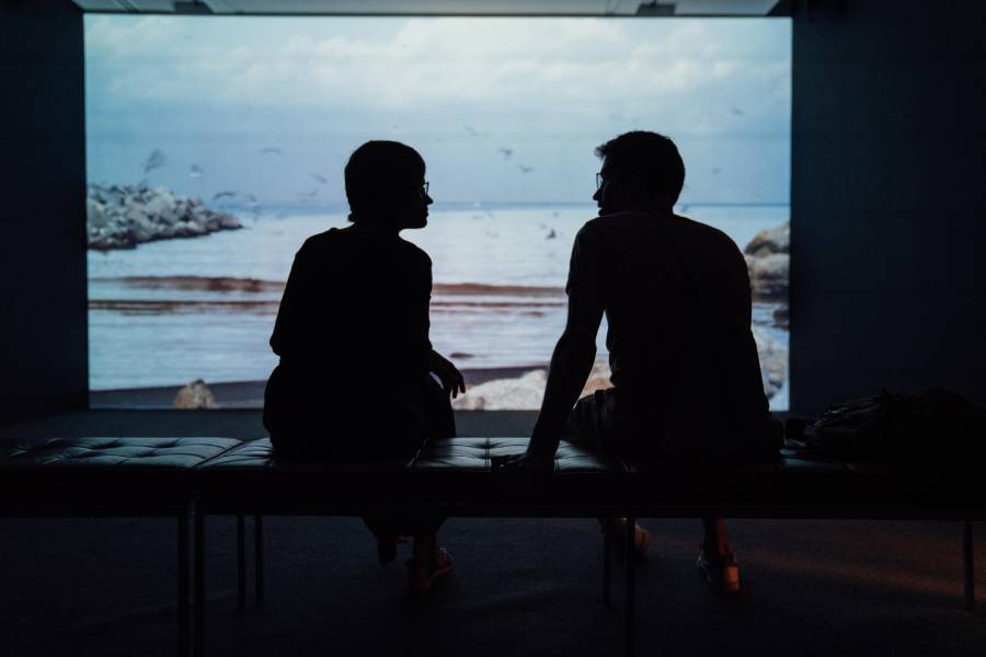 Two backlighted people talking and sitting in front of a projector that shows a video